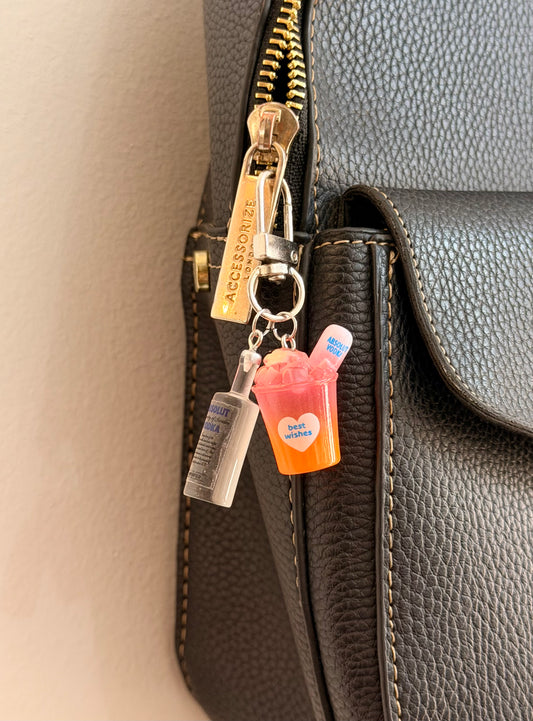 Absolut Orange Cocktail Bag Charm, a miniature clear Absolut vodka bottle with an orange cocktail cup clipped to a black leather handbag zip.