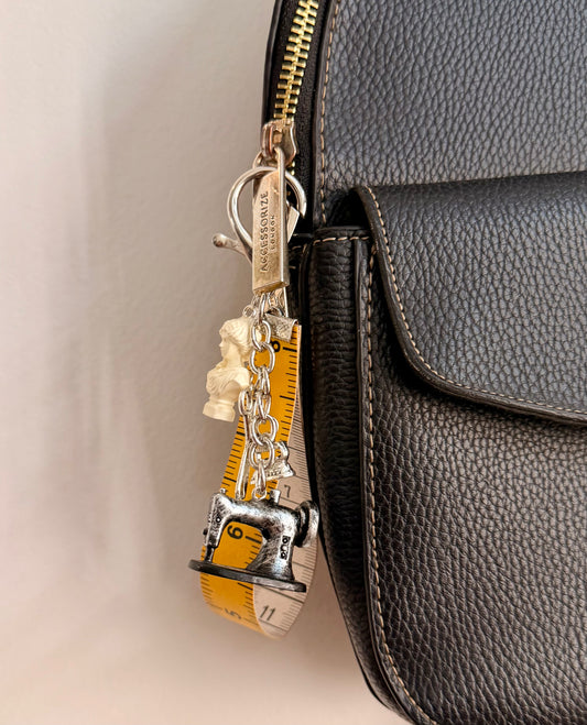 Couture Chaos Bag Charm, a sewing machine, scissors, an iron, a sculpture bust and a measuring tape on a silver chain clipped to a black leather handbag zip.