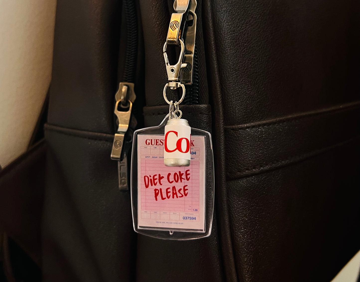 Acrylic keychain with pink receipt reading Diet Coke Please and white mini can on a black bag