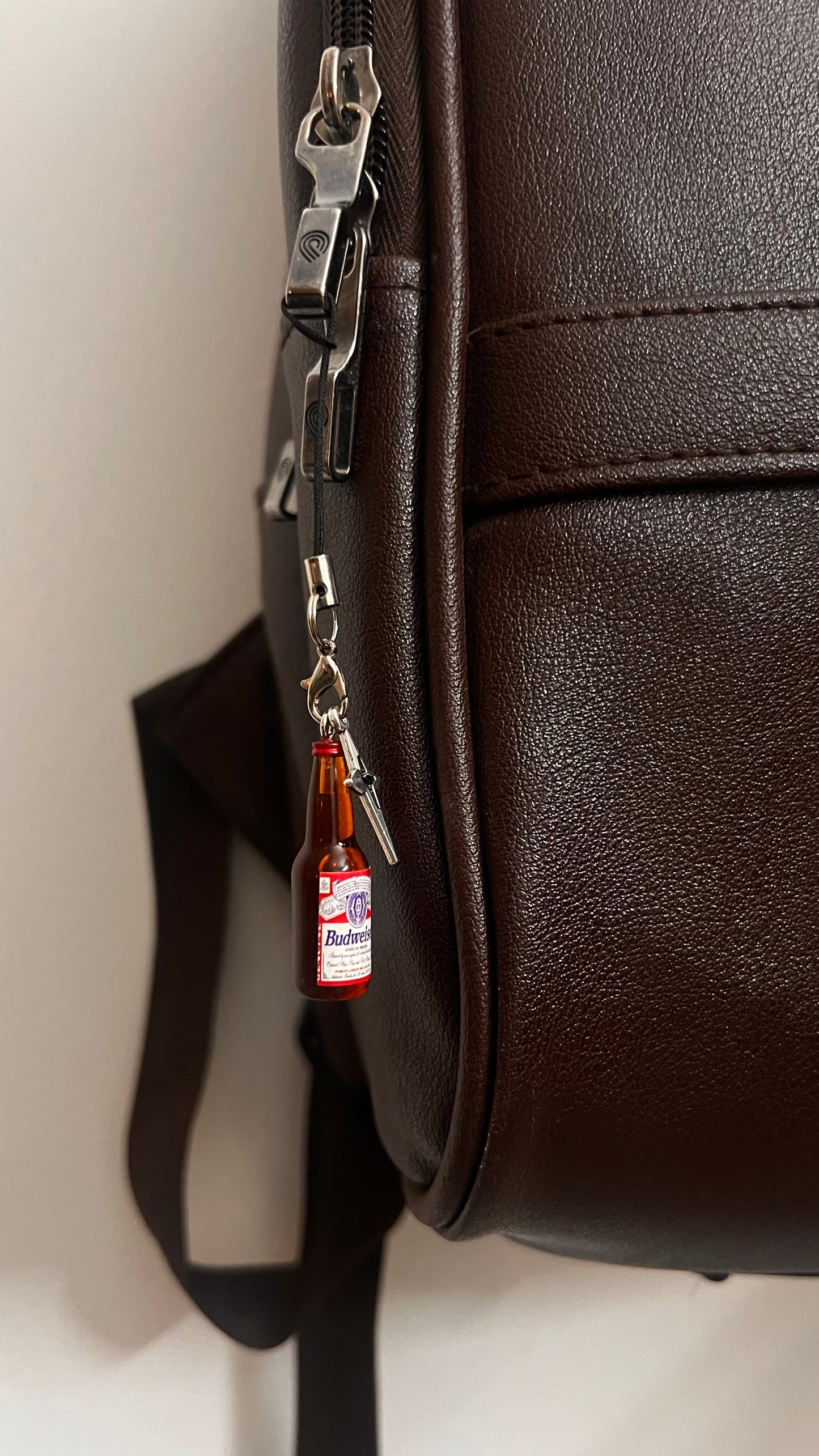 Mini Budweiser bottle phone charm with silver star on a black strap, on a dark brown backpack