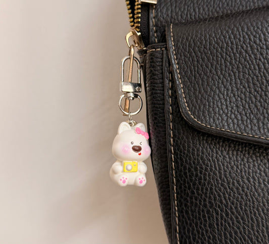 Say Cheese, a white puppy with a pink bow and a yellow camera clipped to a black leather handbag zip.