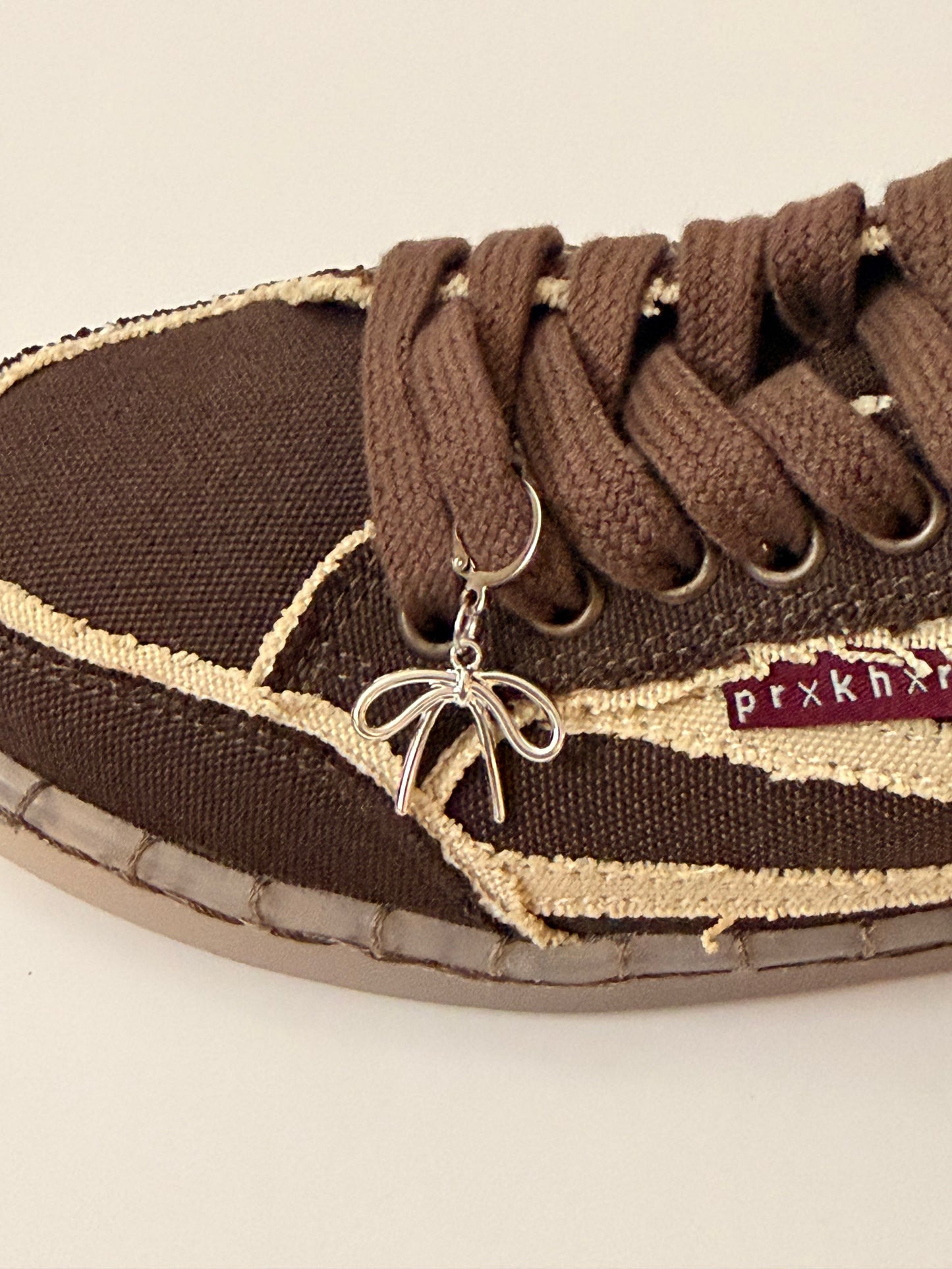 Ribbon Bow Shoe Charm, puffy silver bow charm with wavy tails on the laces of a brown canvas sneaker