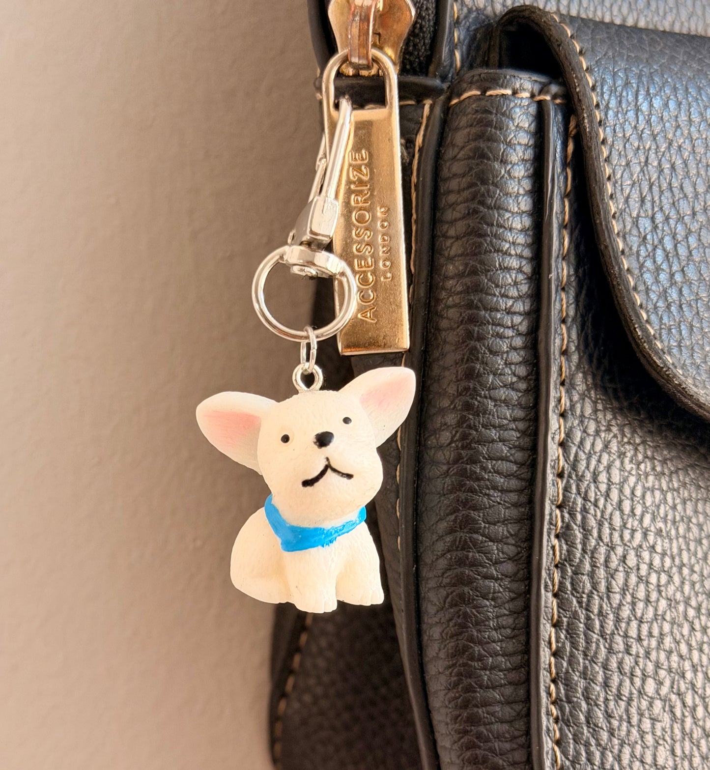 Certified Good Boi, a white french bulldog in a blue collar clipped to a black leather handbag zip.
