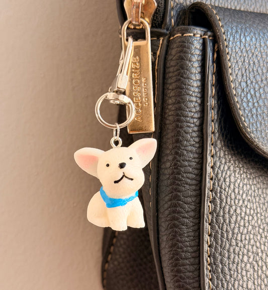 Certified Good Boi, a white french bulldog in a blue collar clipped to a black leather handbag zip.