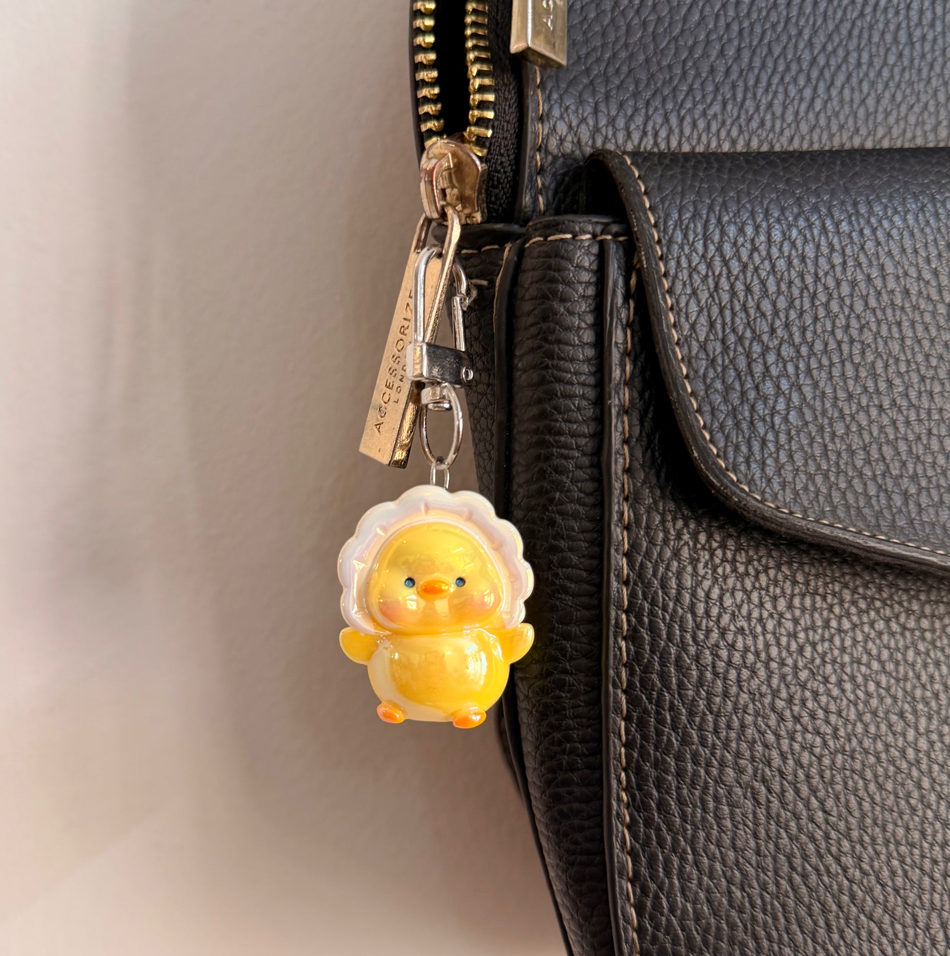 Duckie boo, a yellow duck in a white ruffle collar clipped to a black leather handbag zip.