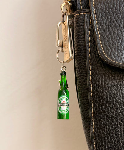 Heineken with Thunderbolt, a miniature green beer bottle with a silver thunderbolt charm clipped to a black leather handbag zip.