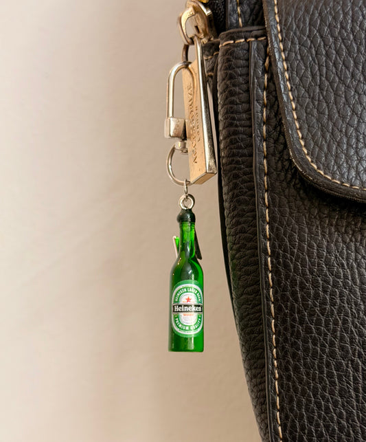 Heineken with Thunderbolt, a miniature green beer bottle with a silver thunderbolt charm clipped to a black leather handbag zip.