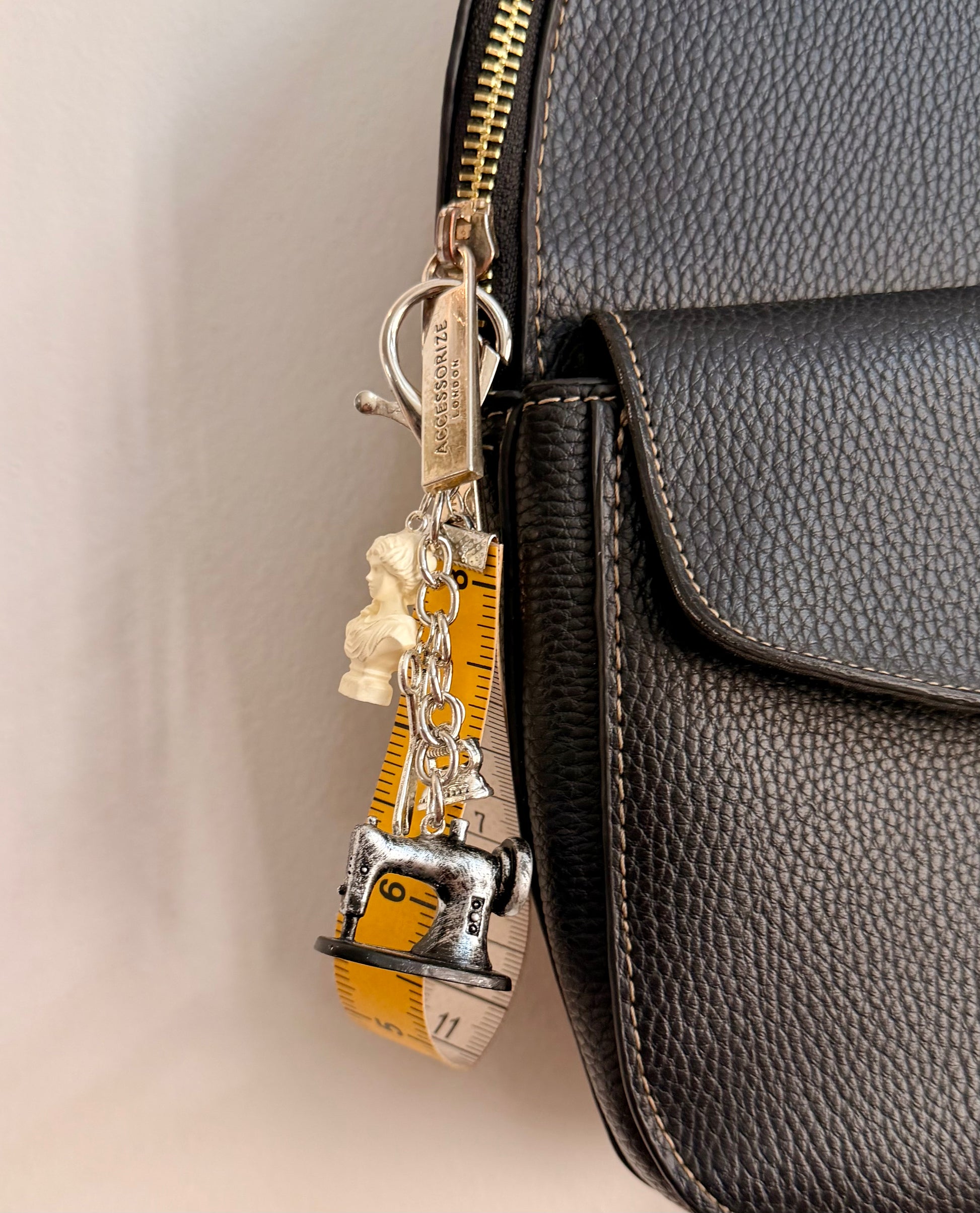 Couture Chaos Bag Charm, a sewing machine, scissors, an iron, a sculpture bust and a measuring tape on a silver chain clipped to a black leather handbag zip.