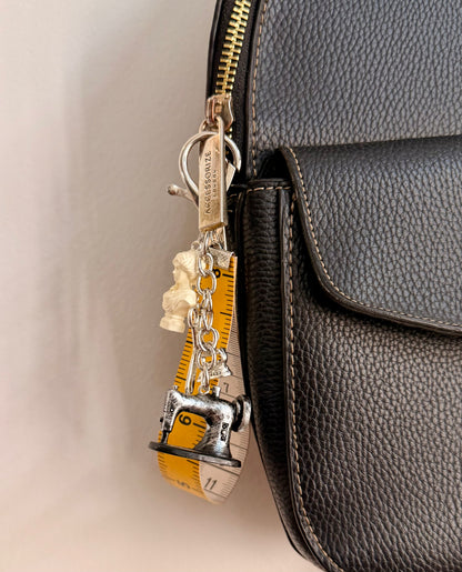 Couture Chaos Bag Charm, a sewing machine, scissors, an iron, a sculpture bust and a measuring tape on a silver chain clipped to a black leather handbag zip.