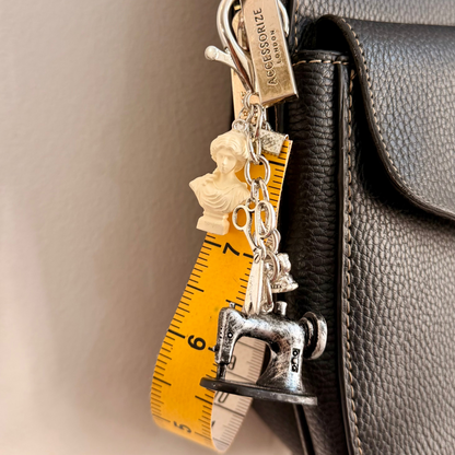 Couture Chaos Bag Charm, a sewing machine, scissors, an iron, a sculpture bust and a measuring tape on a silver chain.