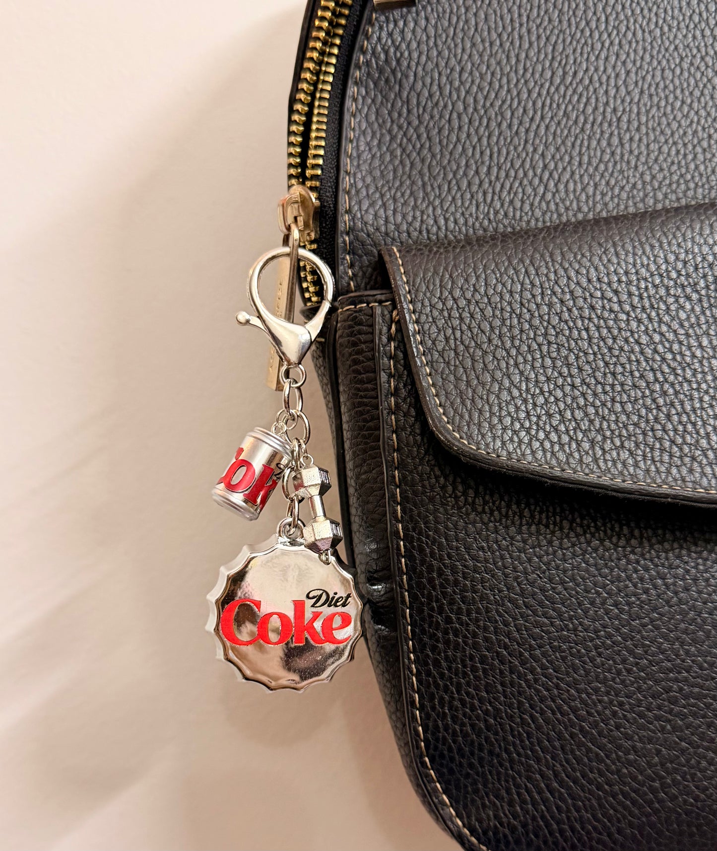 Pop the Cap, an oversized silver Diet Coke bottle cap with a miniature can and a silver dumbbell charm clipped to a black leather handbag zip.