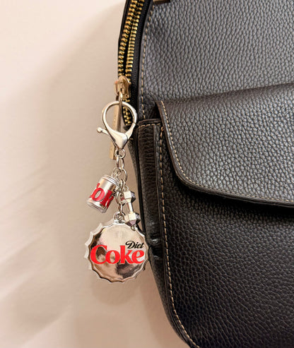 Pop the Cap, an oversized silver Diet Coke bottle cap with a miniature can and a silver dumbbell charm clipped to a black leather handbag zip.