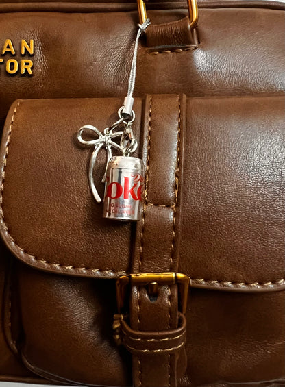 Metallic Diet Coke can charm with silver ribbon bow hanging on a brown leather satchel