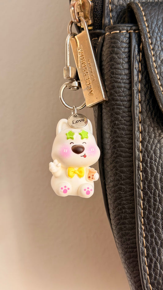 In my Boba Era, a white puppy in green star sunglasses holding a boba cup clipped to a black leather handbag zip.