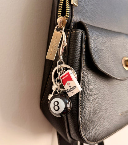 Lucky 8, a black 8 ball and a miniature cigarette pack on a silver keyring clipped to a black leather handbag zip.