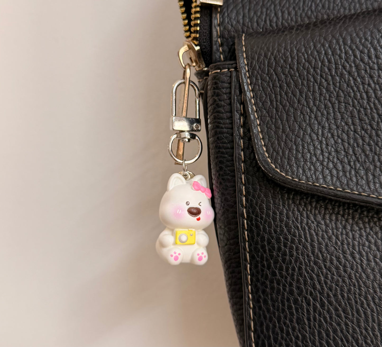 Say Cheese, a white puppy with a pink bow and a yellow camera clipped to a black leather handbag zip.
