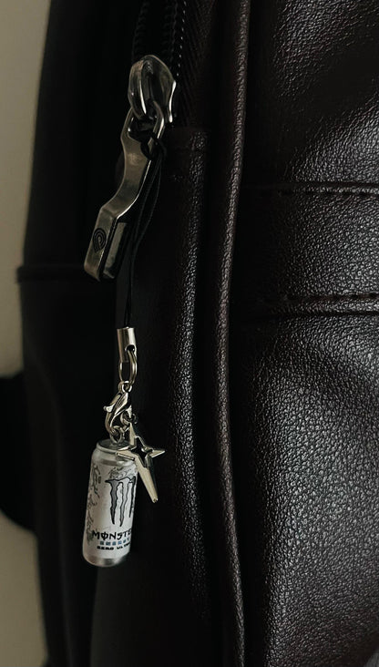 White Monster Energy can charm with star pendant on a black phone strap, dark brown bag