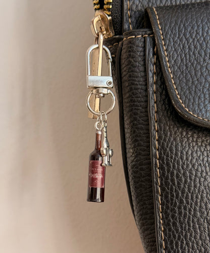 Wine O'Clock, a miniature red wine bottle with a silver corkscrew clipped to a black leather handbag zip.